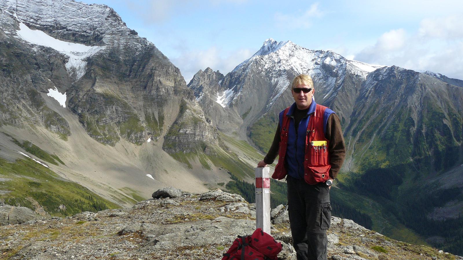 A man standing at a claim post with mountains in the background